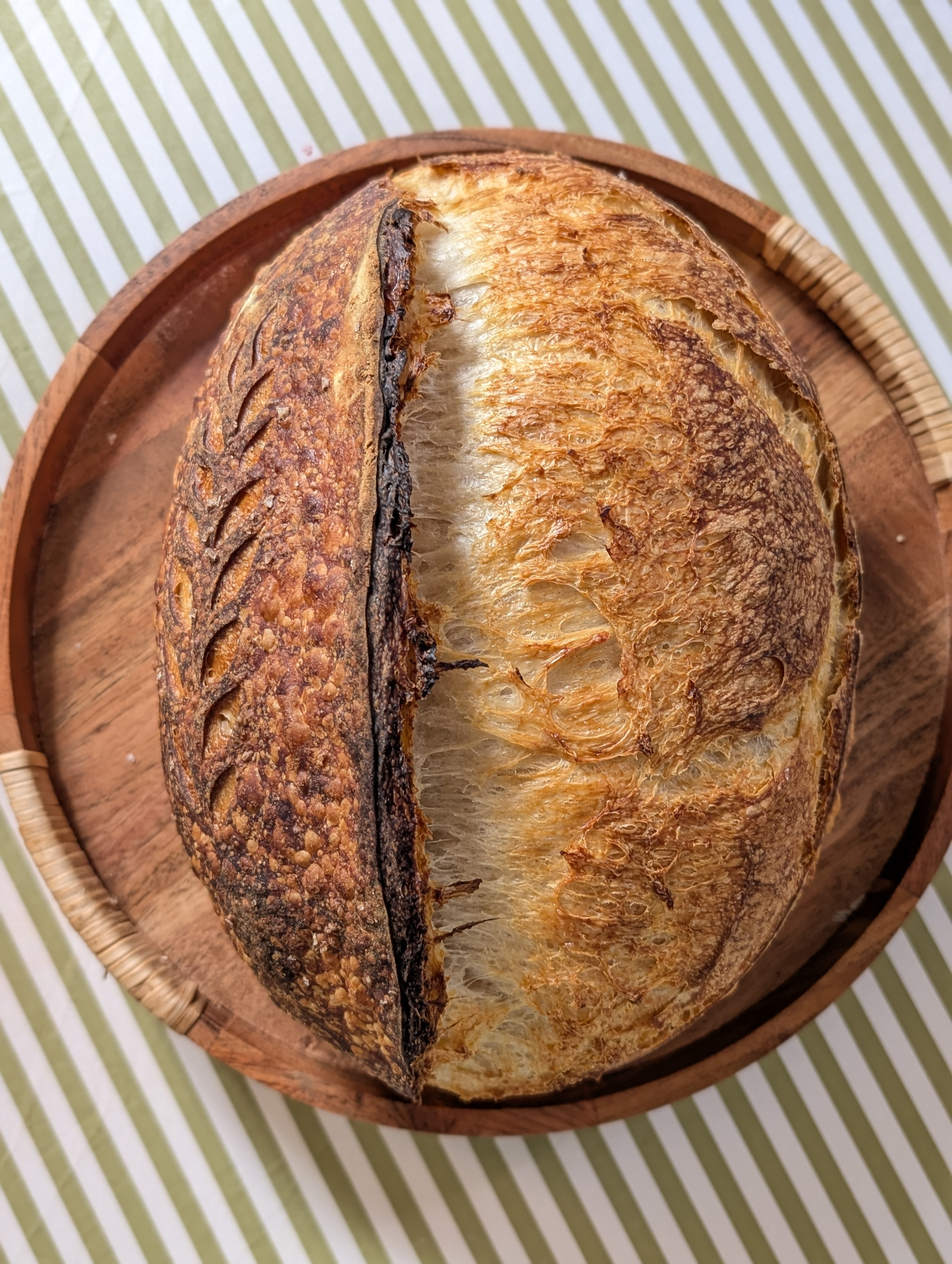A scored oval sourdough loaf with a dark blistered crust resting on a round wooden tray